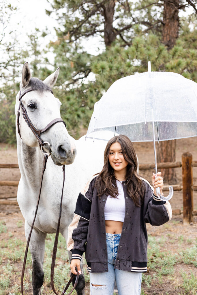 A young woman and her horse stand next to each other in a field as it's raining and the young woman is holding a clear umbrella.