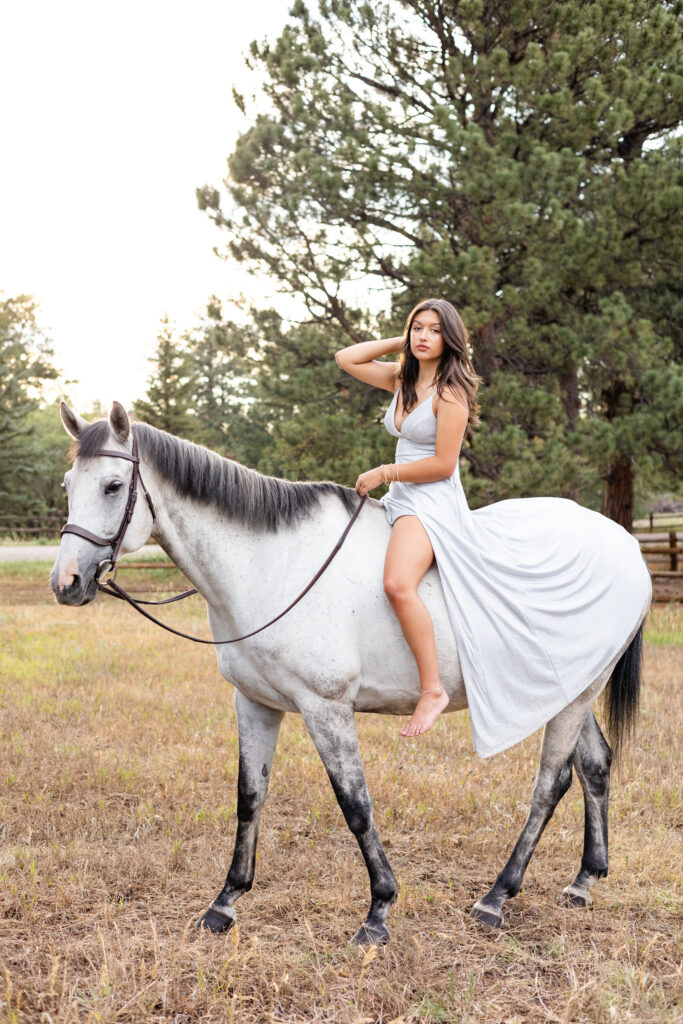 A young woman in a silver dress sits on her white horse and runs one had through her hair as she looks at the camera.