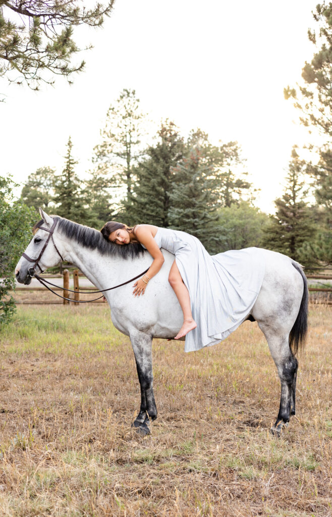 A young woman in a silver dress lays on her white horse's back with her face turned toward the camera.