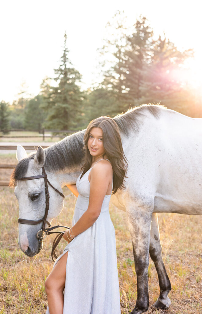 A young woman in a silver dress stands in front of her white horse and smiles at the camera.