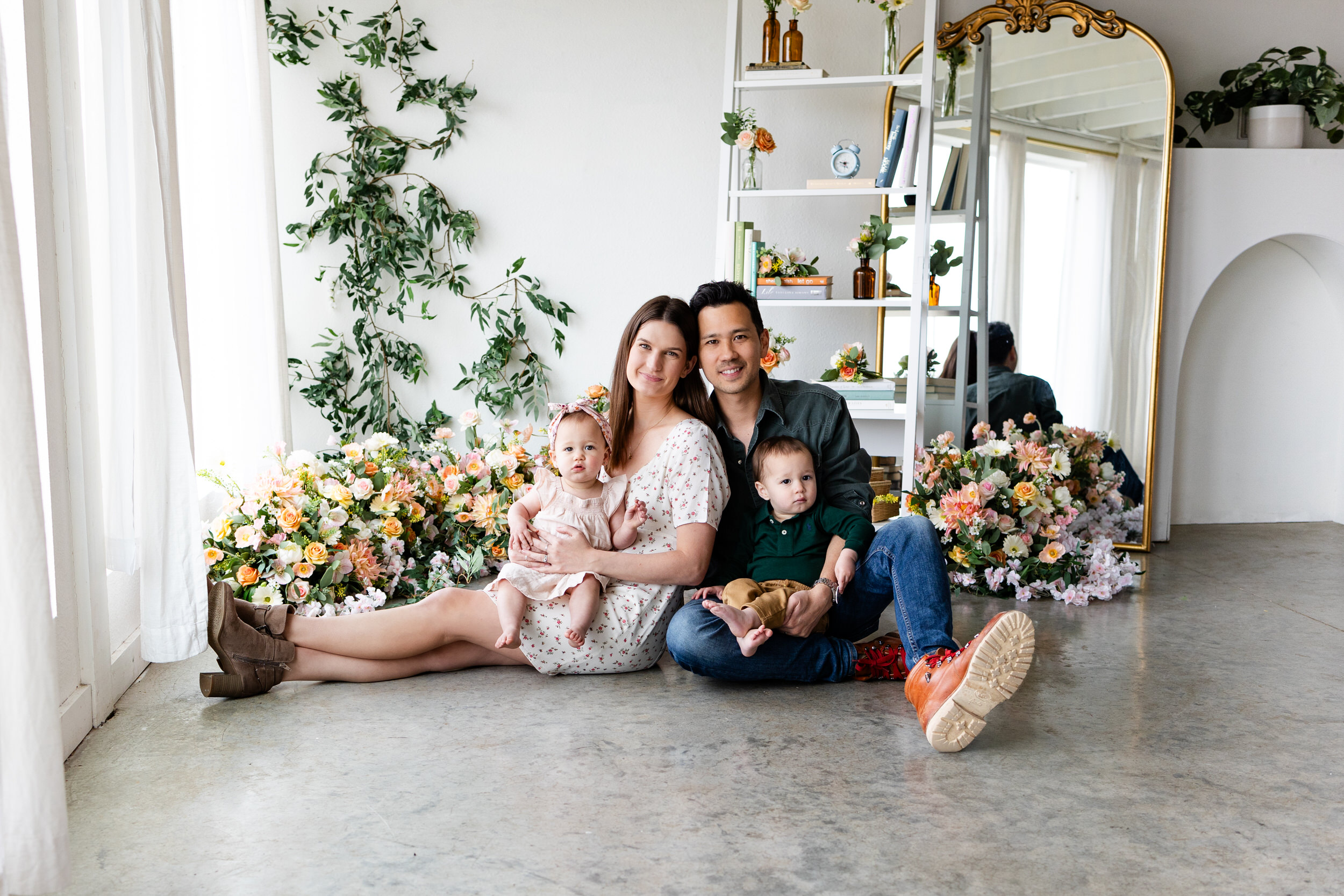 A mom and dad sit on the floor of a studio filled with flowers with each one holding one of their twins on their lap.