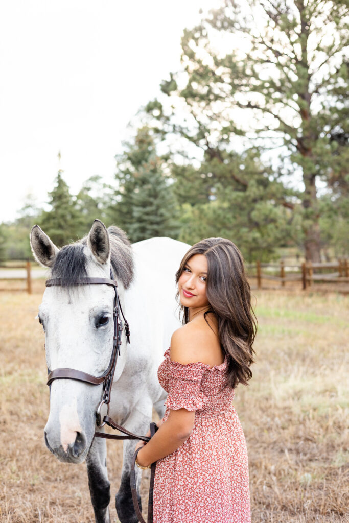 A young woman in a pink floral dress stands with her back to her the camera and her horse at her side as she looks back over her shoulder and smiles at the camera. 