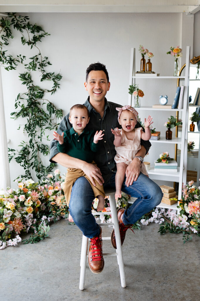 Dad sits on a stool in a studio filled with flowers holding both of his twins who are smiling and clapping.