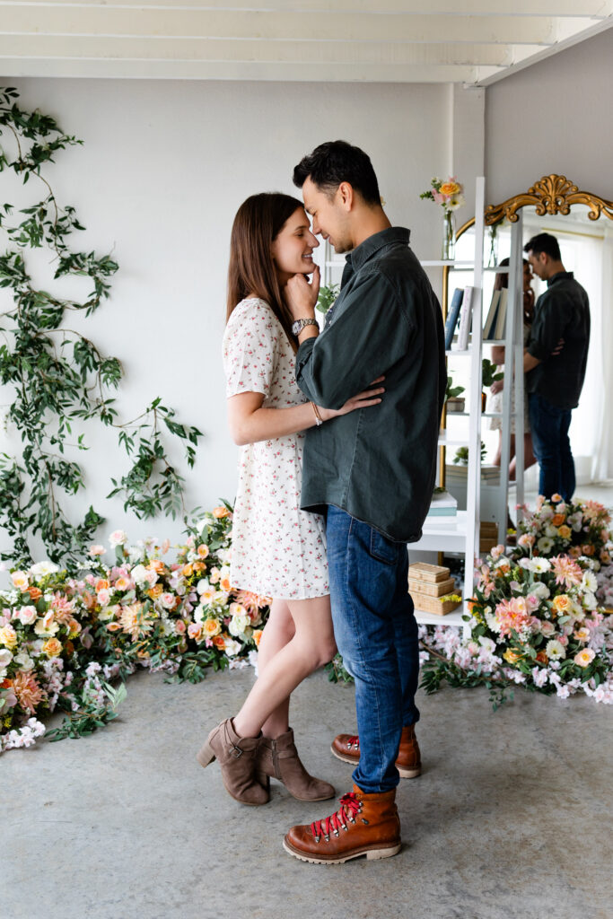 Husband and wife stand forehead to forehead in a studio filled with flowers.