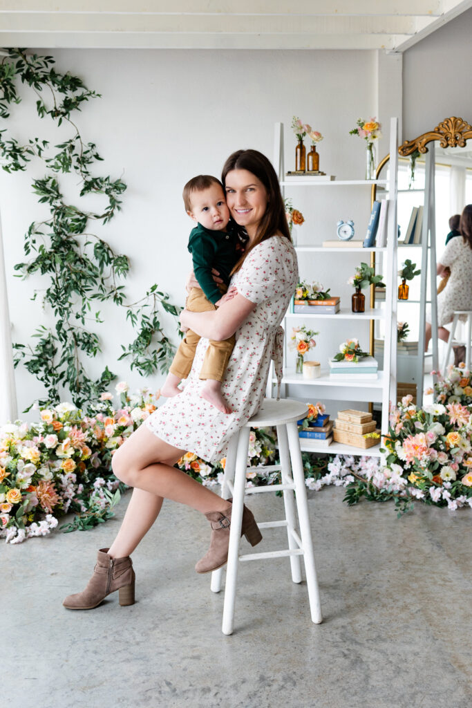 A mom sits on a stool in a studio filled with flowers and holds her young son on her lap.