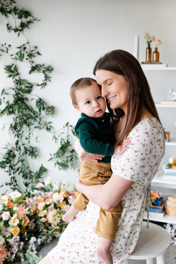 A mom sits on a stool in a studio filled with flowers and holds her young son on her lap.