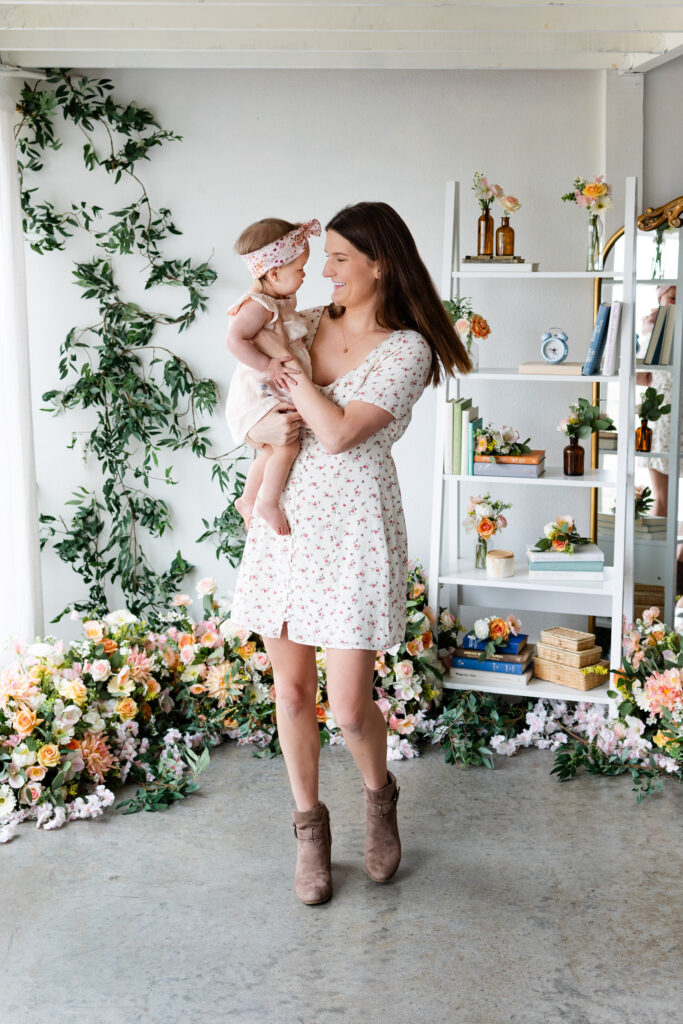 A mom spins around with her young daughter in her arms in a studio filled with flowers.