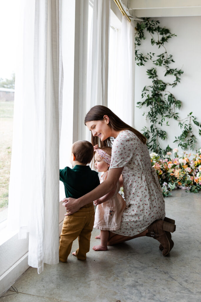 Two young twins look out a window as their mom sits near them with her arms around them.