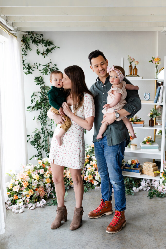 A mom and dad each hold one of their twins while mom kisses her son's cheek and dad smiles at his daughter.