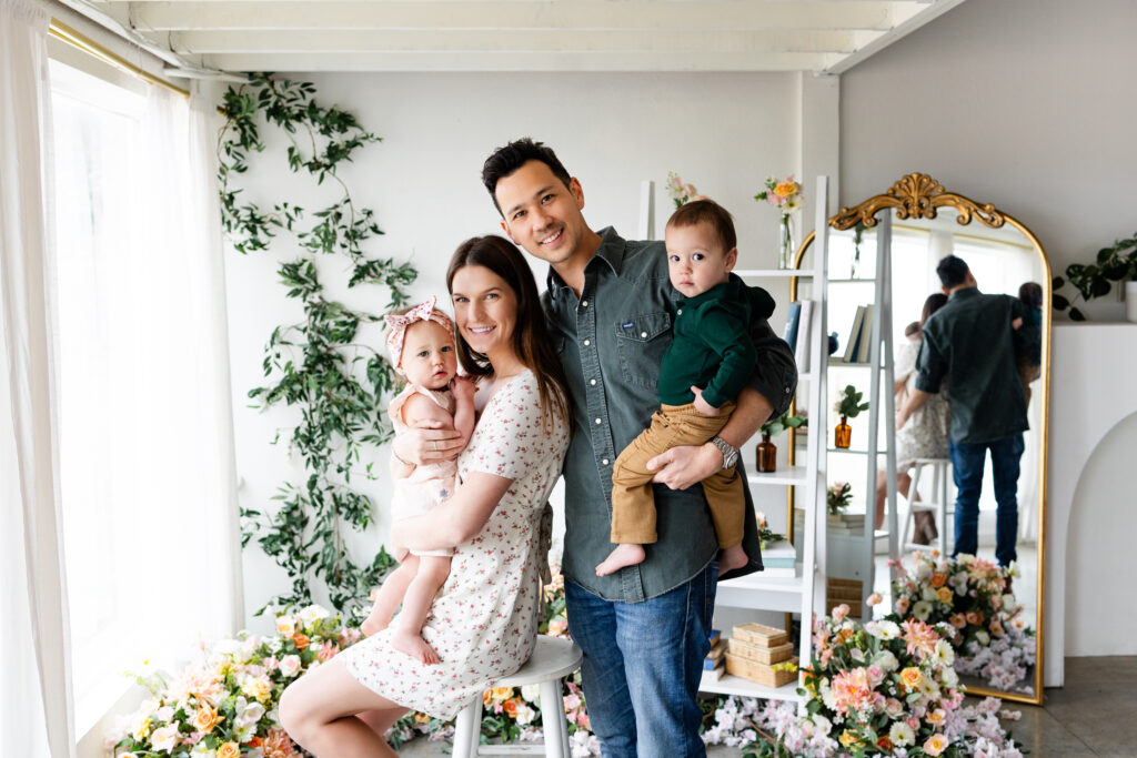 A family of four smiles at the camera in a studio filled with flowers.