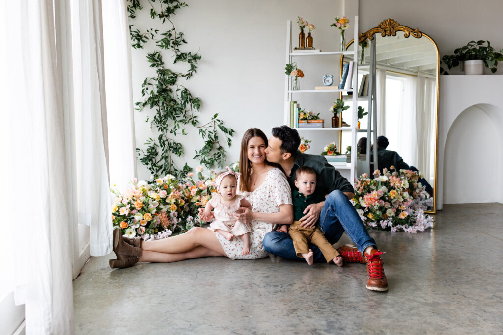 Dad kisses mom's cheek as they both sit on the floor of a studio filled with flowers with each parent holding one of their young twins in their lap.