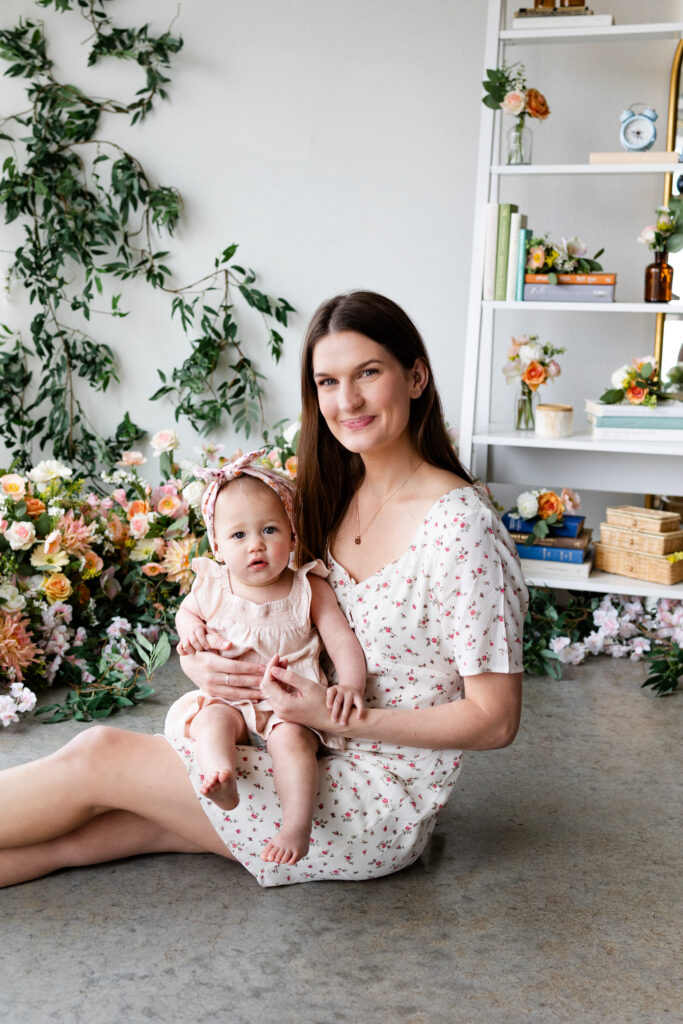 Mom sits on the floor of a studio filled with flowers with her young daughter on her lap.