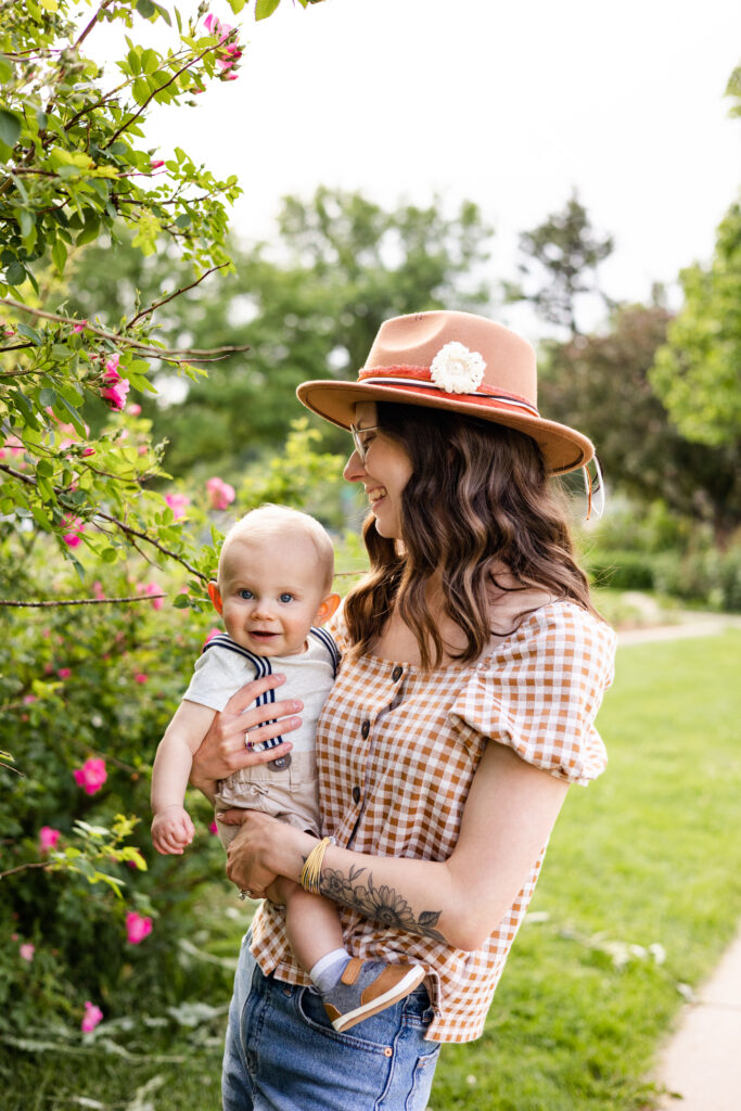 A mom holds and smiles at her baby boy in front of a rose bush filled with pink roses.