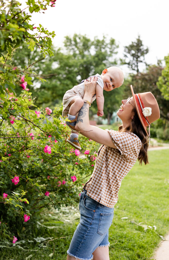 A mom holds her baby boy up in the air as he smiles at the camera with a pink rose bush in the background.