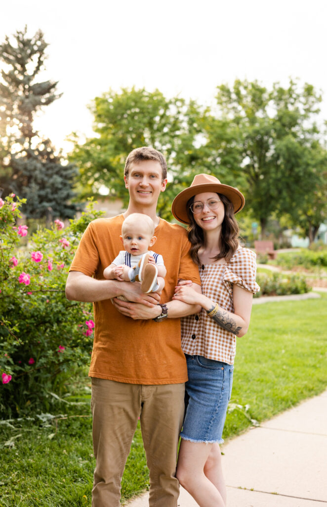 A mom and dad hold their baby boy and they all smile at the camera with a pink rose bush in the background.