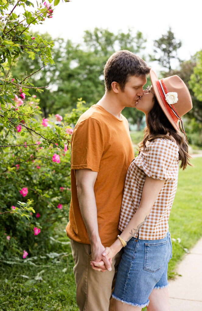 A husband and wife share a kiss in front of a rose bush filled with pink roses.