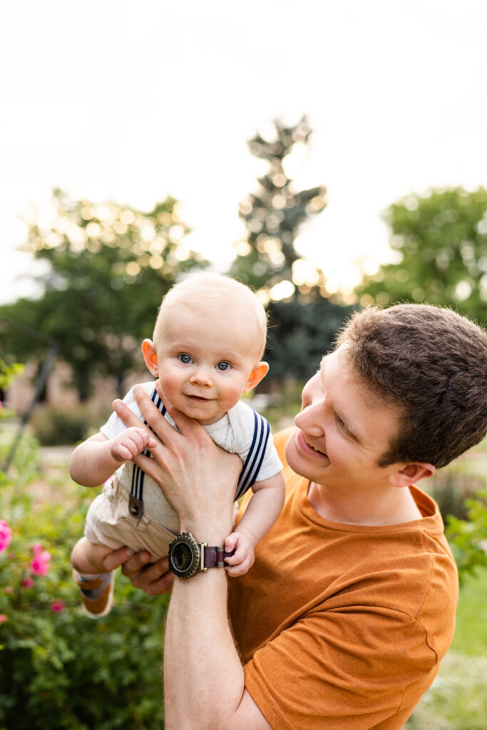 A dad holds his baby boy up in the air and looks at him while his baby looks at the camera.