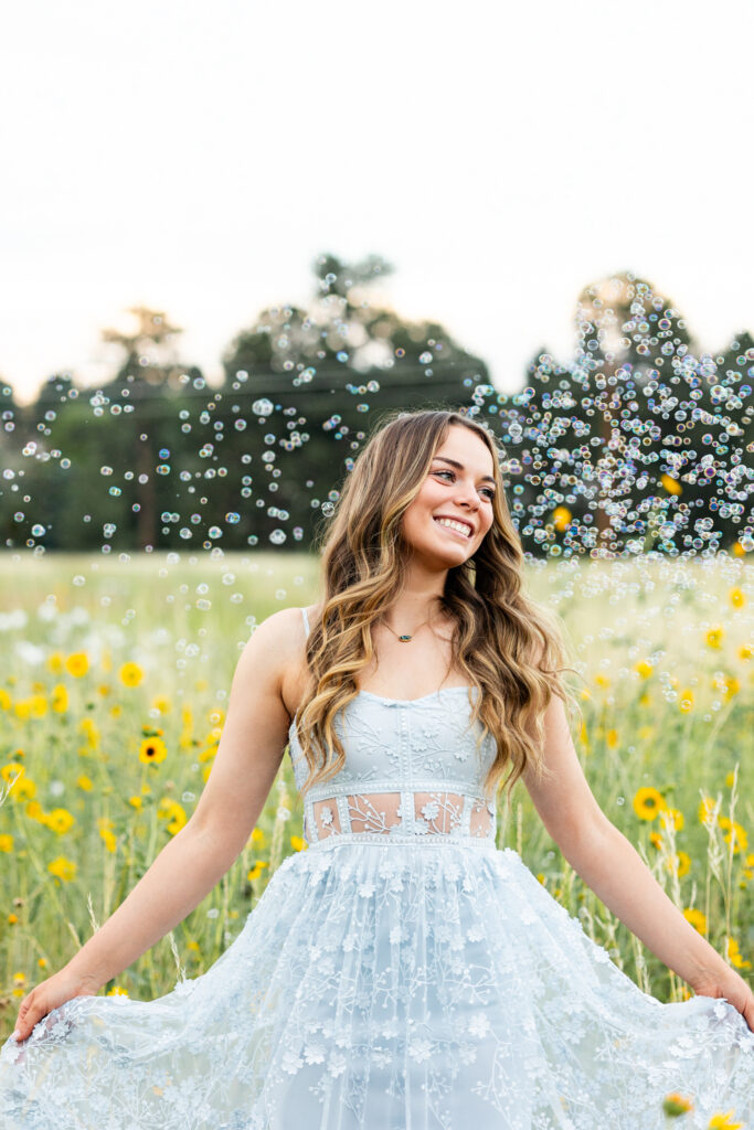 A young woman in a blue dress stands in a field of yellow wildflowers holding onto her skirt and is surrounded by bubbles.