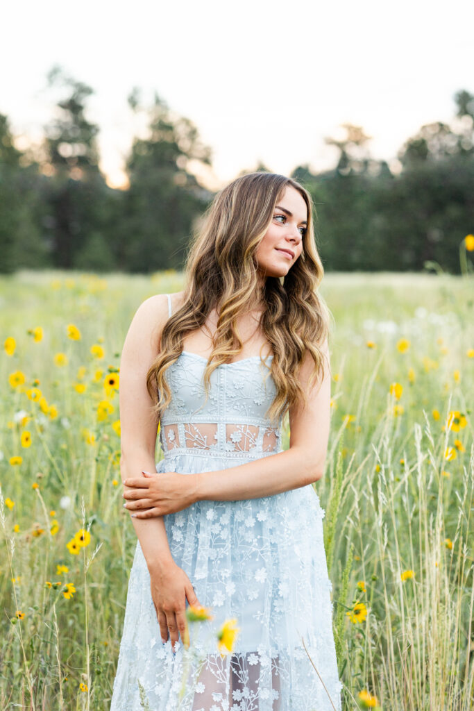 A young woman in a blue dress stands in a field of yellow wildflowers and looks to her left over her shoulder.