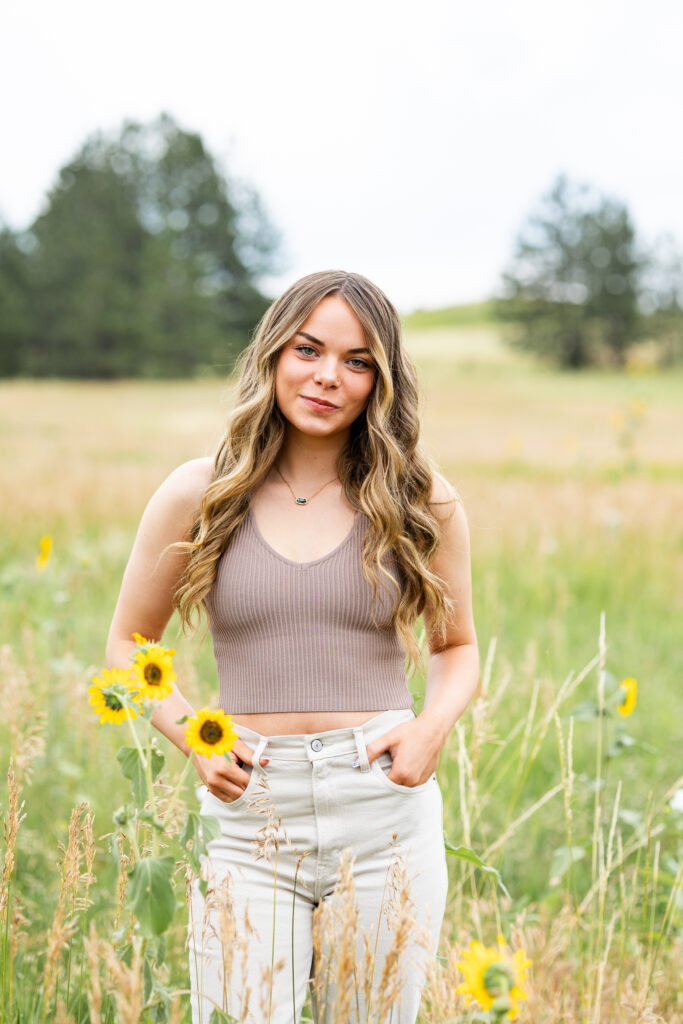 A young woman in a tan crop top and white pants stands in a field of yellow wildflowers and smiles at the camera. 