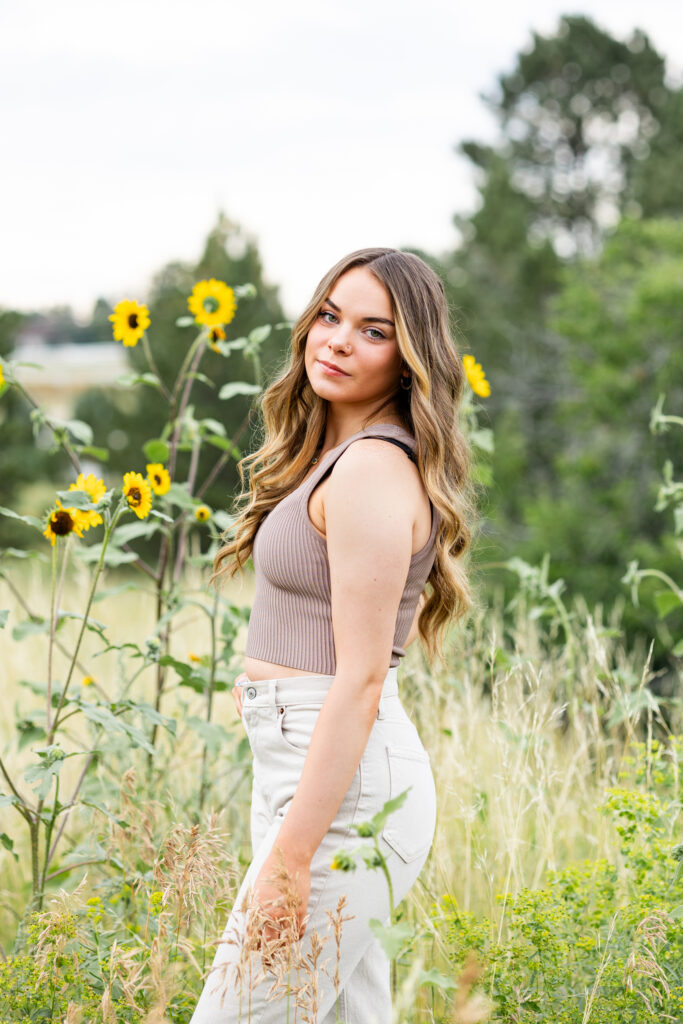 A young woman in a tan crop top and white pants stands with her left side toward the camera and she looks over her left shoulder at the camera.