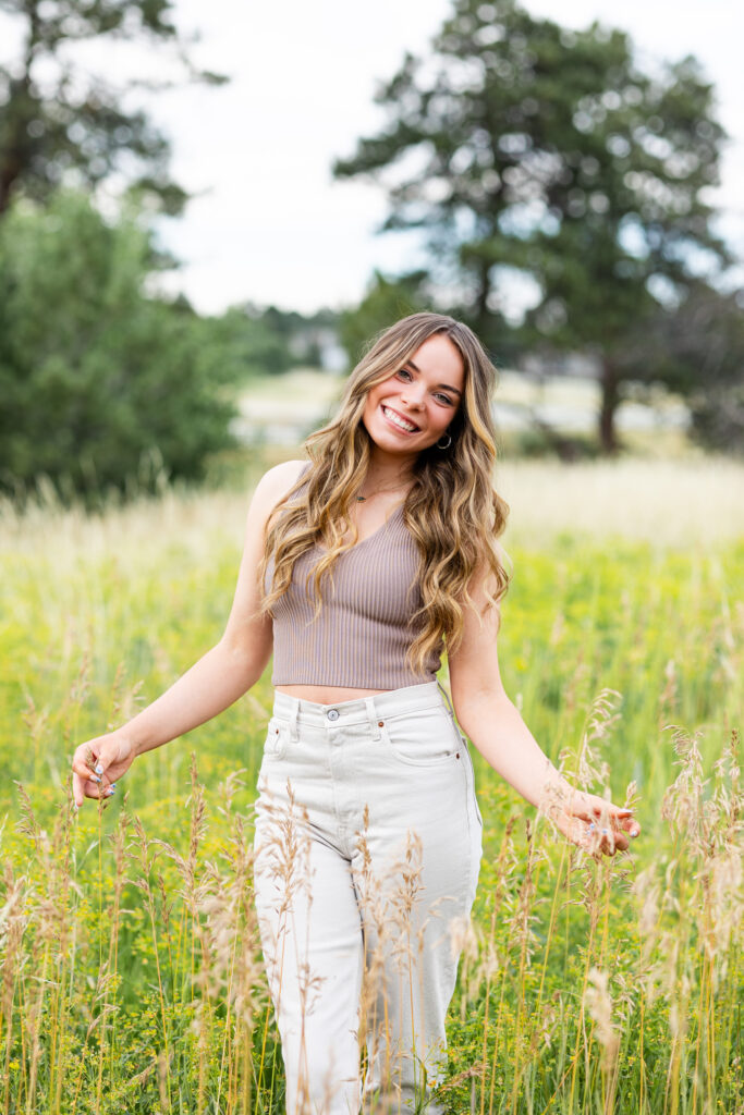 A young woman in a tan crop top and white pants stands in a field of tall grass and smiles at the camera. 