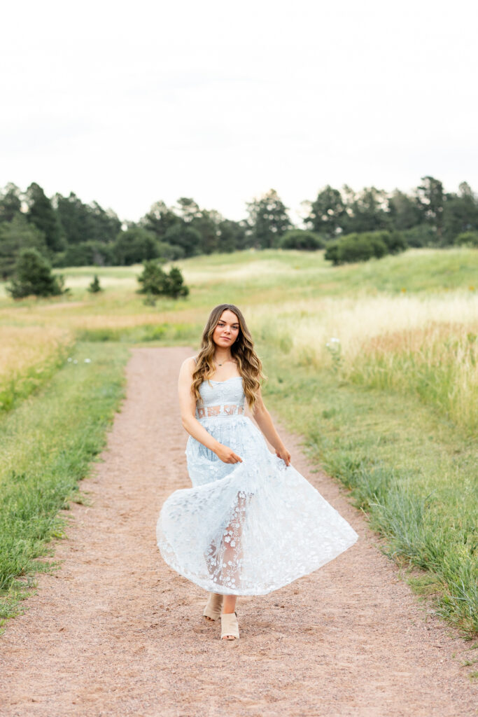 A young woman in a blue dress swishes her skirt back and forth as she walks down a dirt path toward the camera.