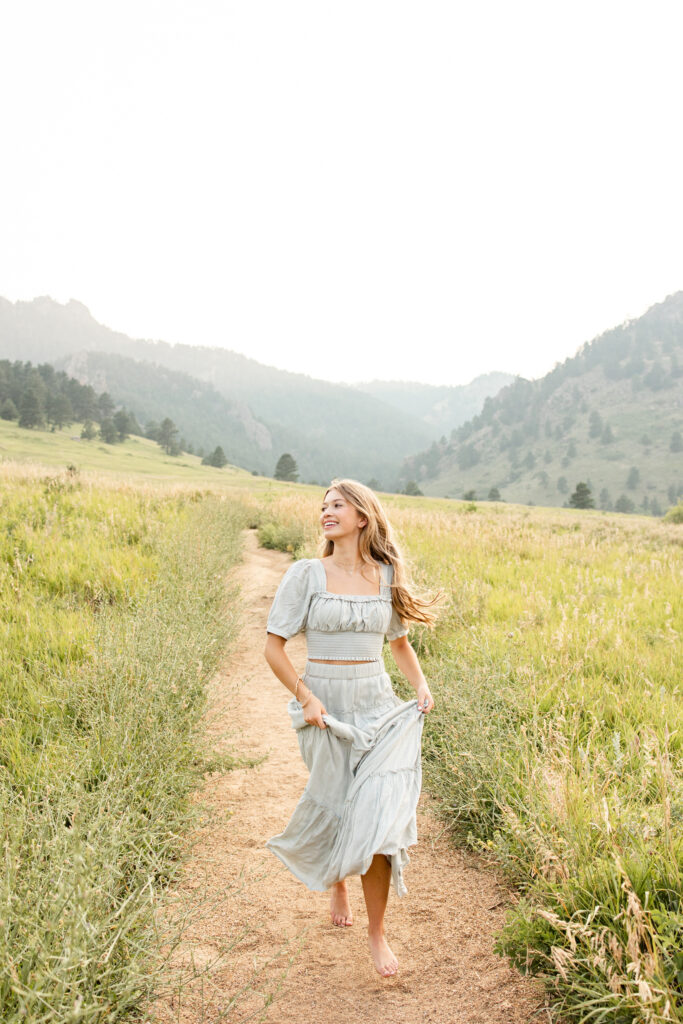 A young woman in a sage green dress runs down a mountain path barefoot.