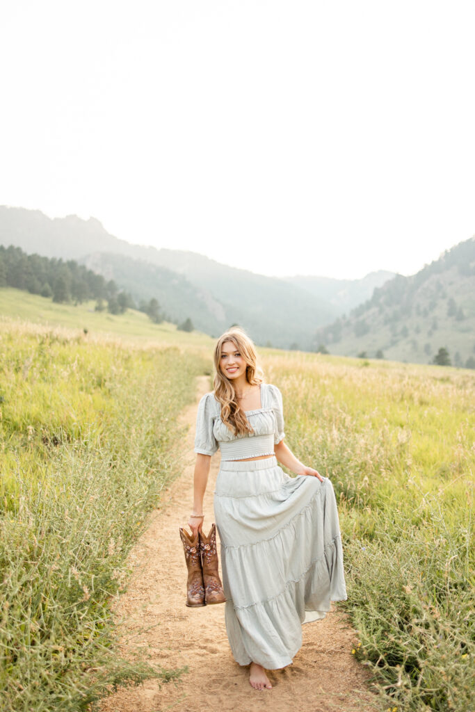 A young woman in a sage green dress carries cowgirl boots in one hand and holds her skirt in the other as she walks toward the camera.