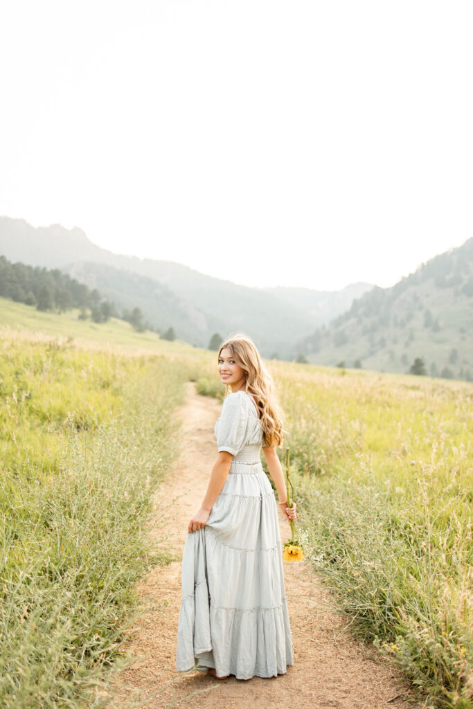 A young woman in a sage green dress faces away from the camera and looks back over her shoulder at the camera.