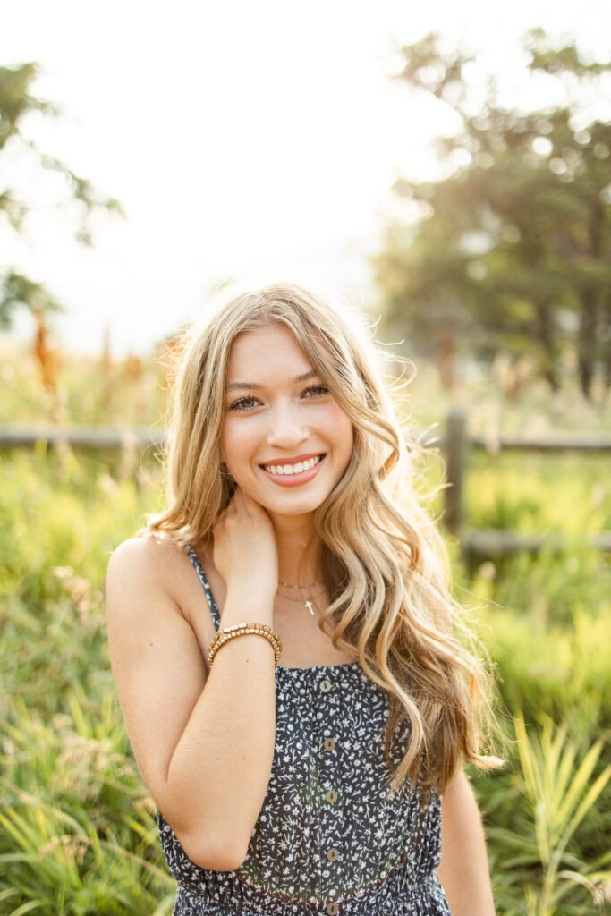 A young woman in a navy blue dress smiles at the camera with one hand in her hair. 