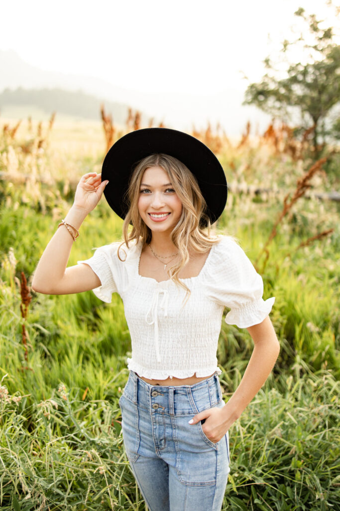 A young woman smiles at the camera and holds the brim of the hat she is wearing with one hand.