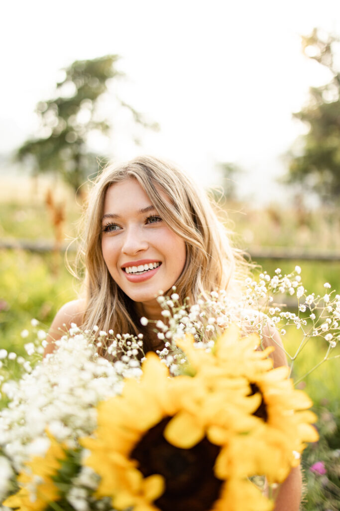 A young woman holding a bouquet of sunflowers and baby's breath looks to her right and smiles.