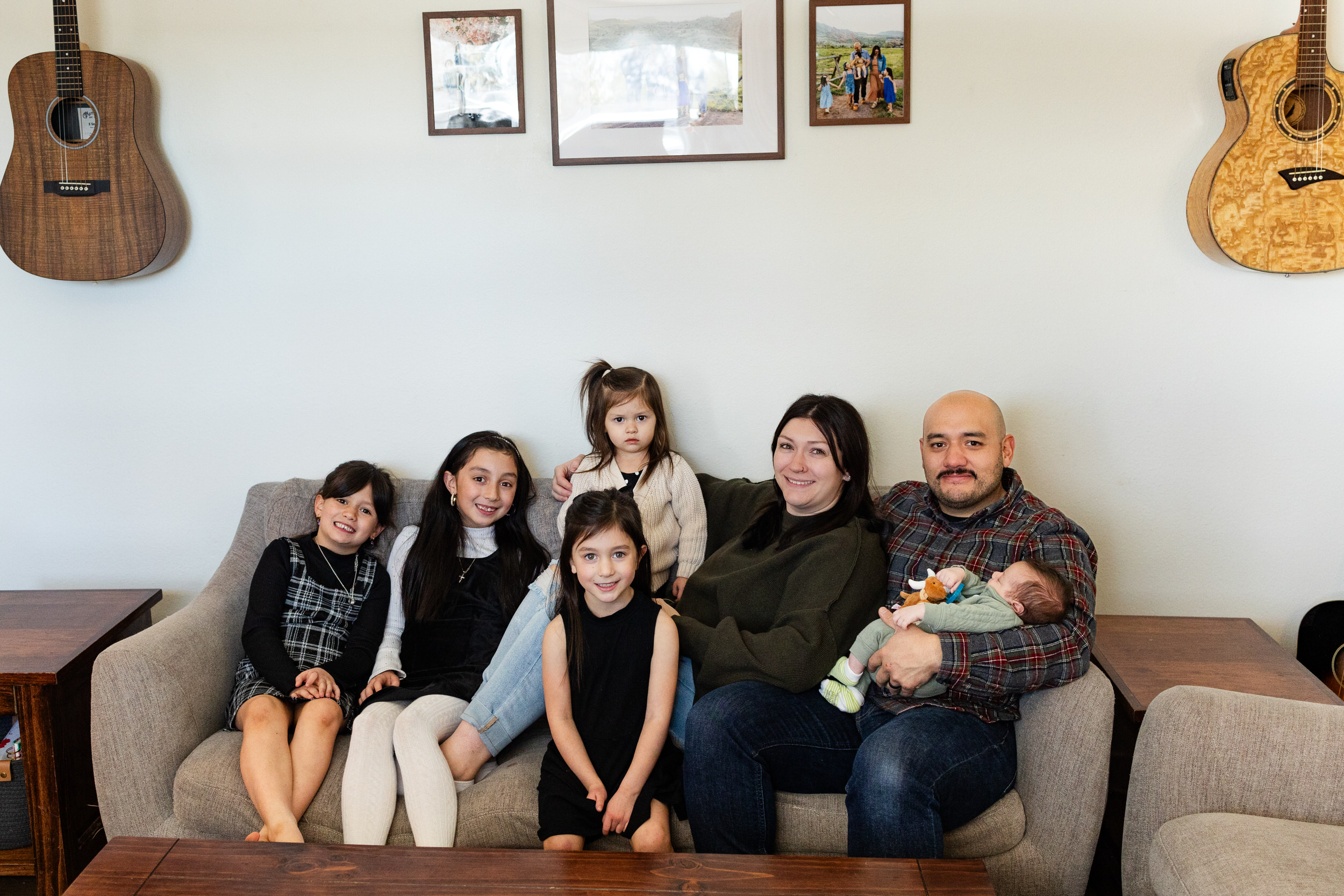A family of seven sits on their couch and smiles at the camera.