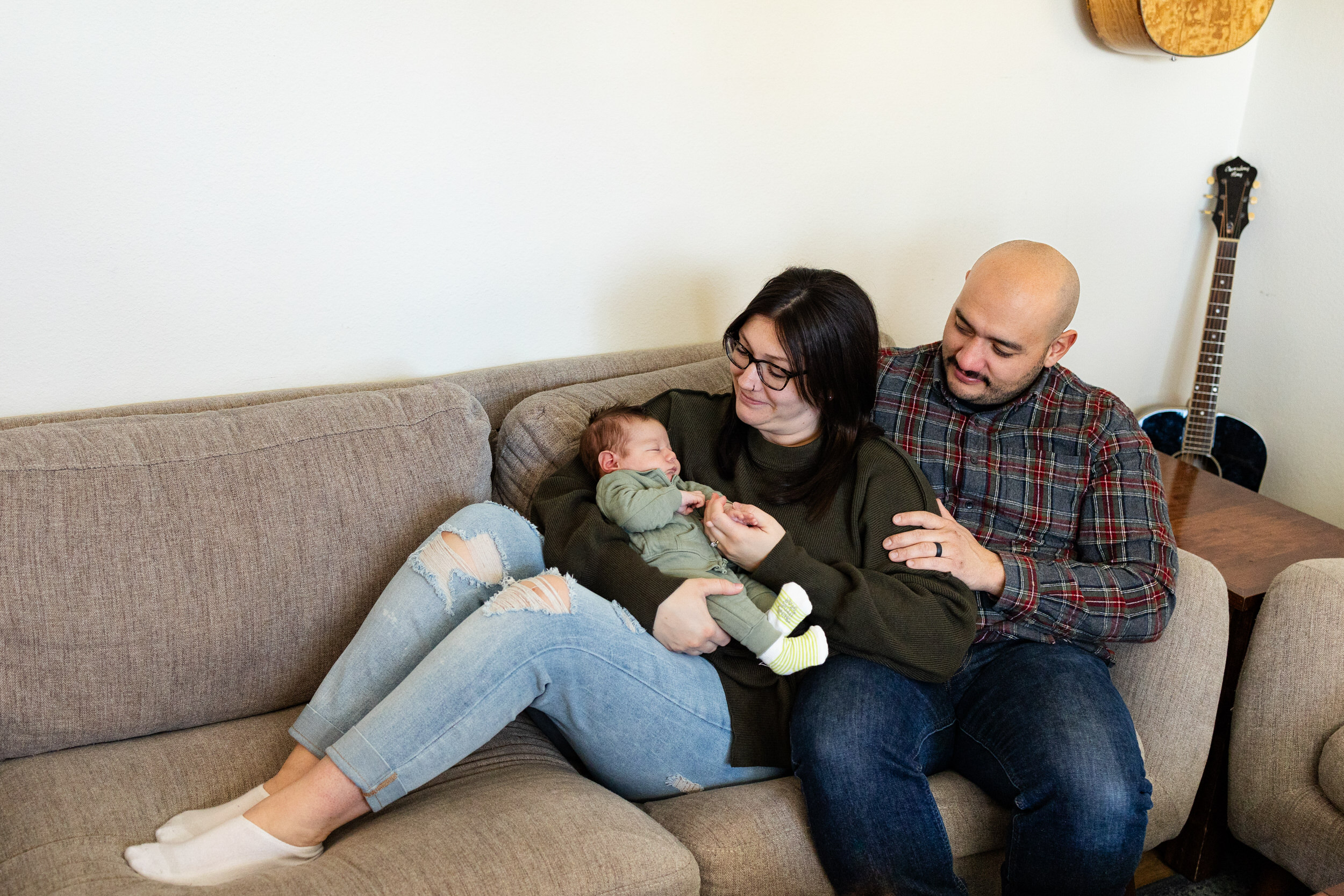 A mom and dad snuggle on the couch while holding their newborn son.