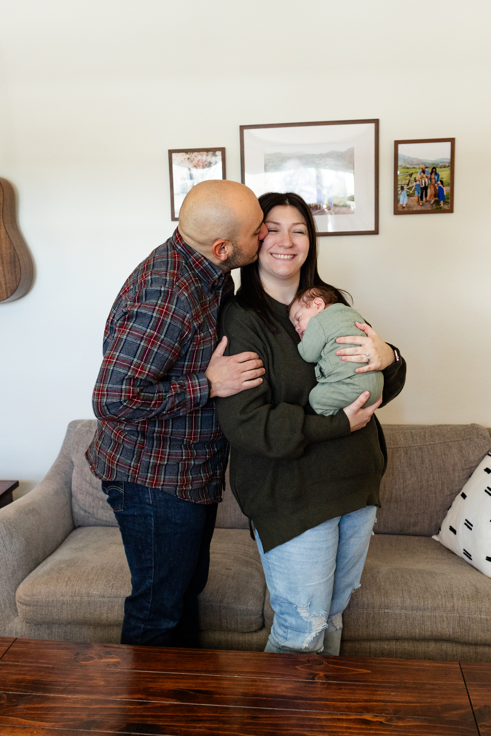 Dad kisses mom's cheek as she smiles and holds newborn baby boy against her chest.