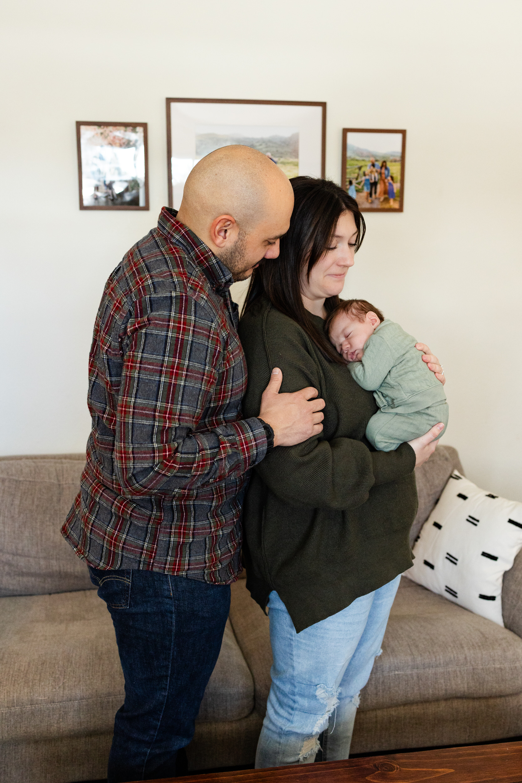 Mom and dad snuggle each other and smile while mom holds sleeping newborn baby boy against her chest.