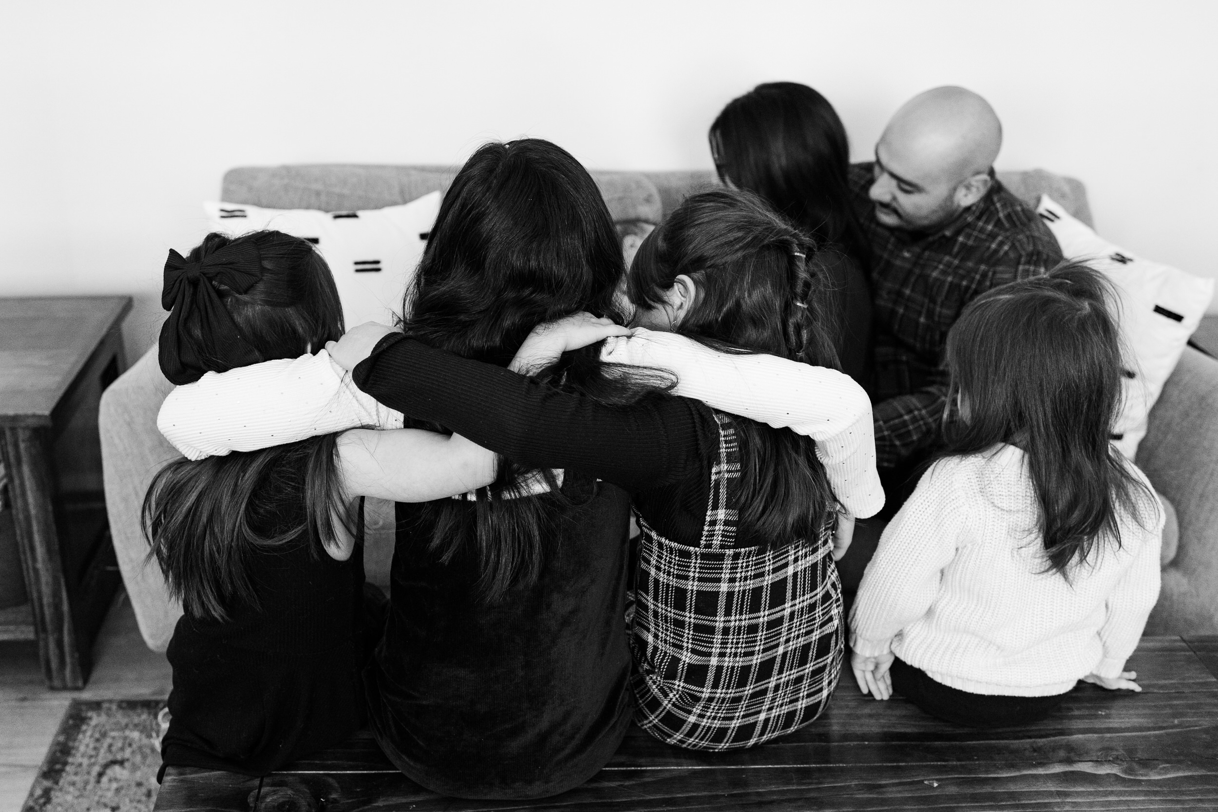 Four young sisters sit on a coffee table in their living room with their arms around each other's shoulders while mom and dad snuggle their newborn baby brother on the couch in the background.