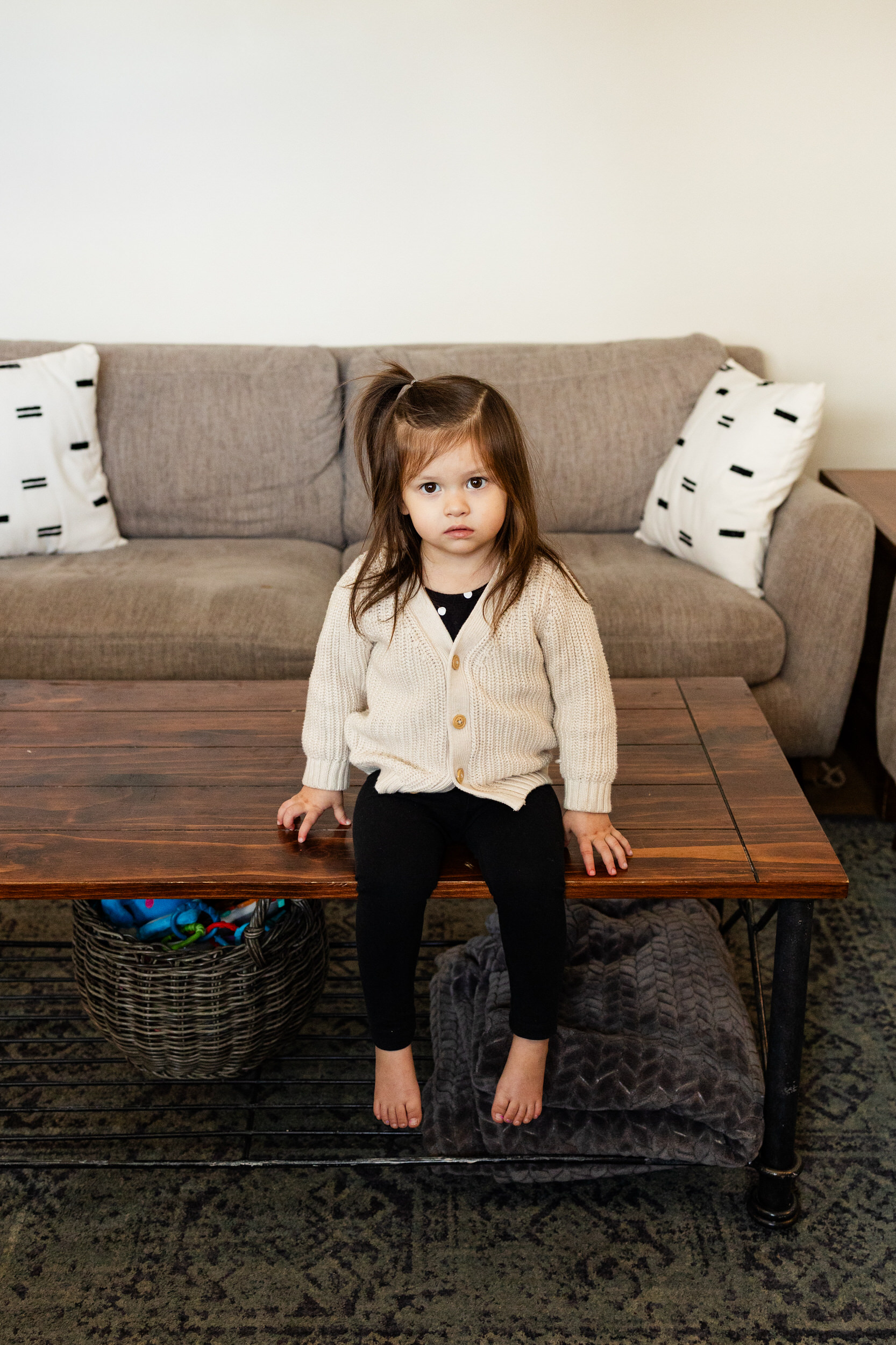 A young girl sits on a coffee table in her living room and looks at the camera.