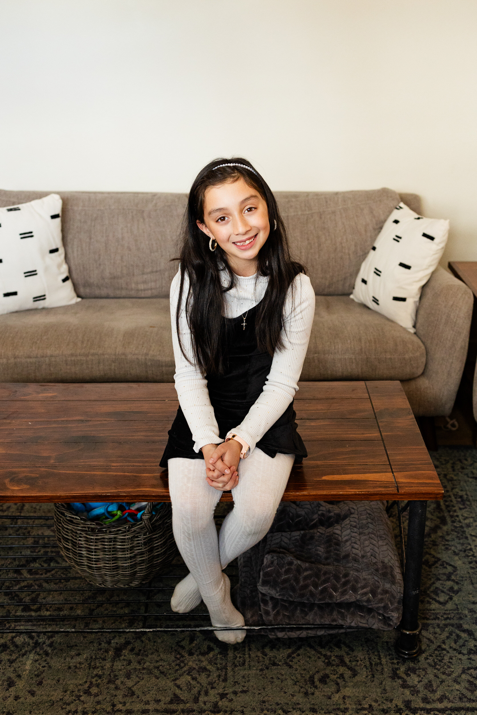 A young girl sits on a coffee table in her living room and smiles at the camera.