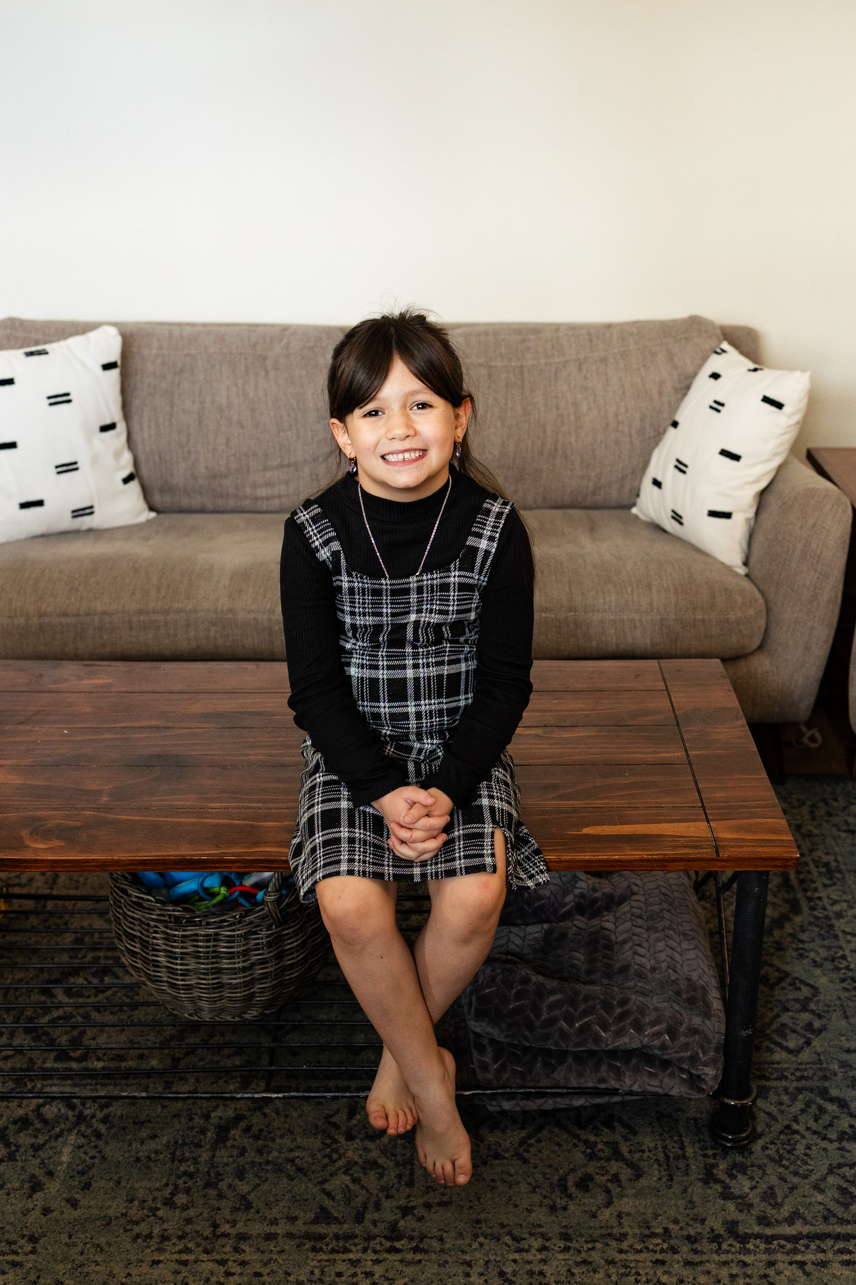 A young girl sits on a coffee table in her living room and smiles at the camera.
