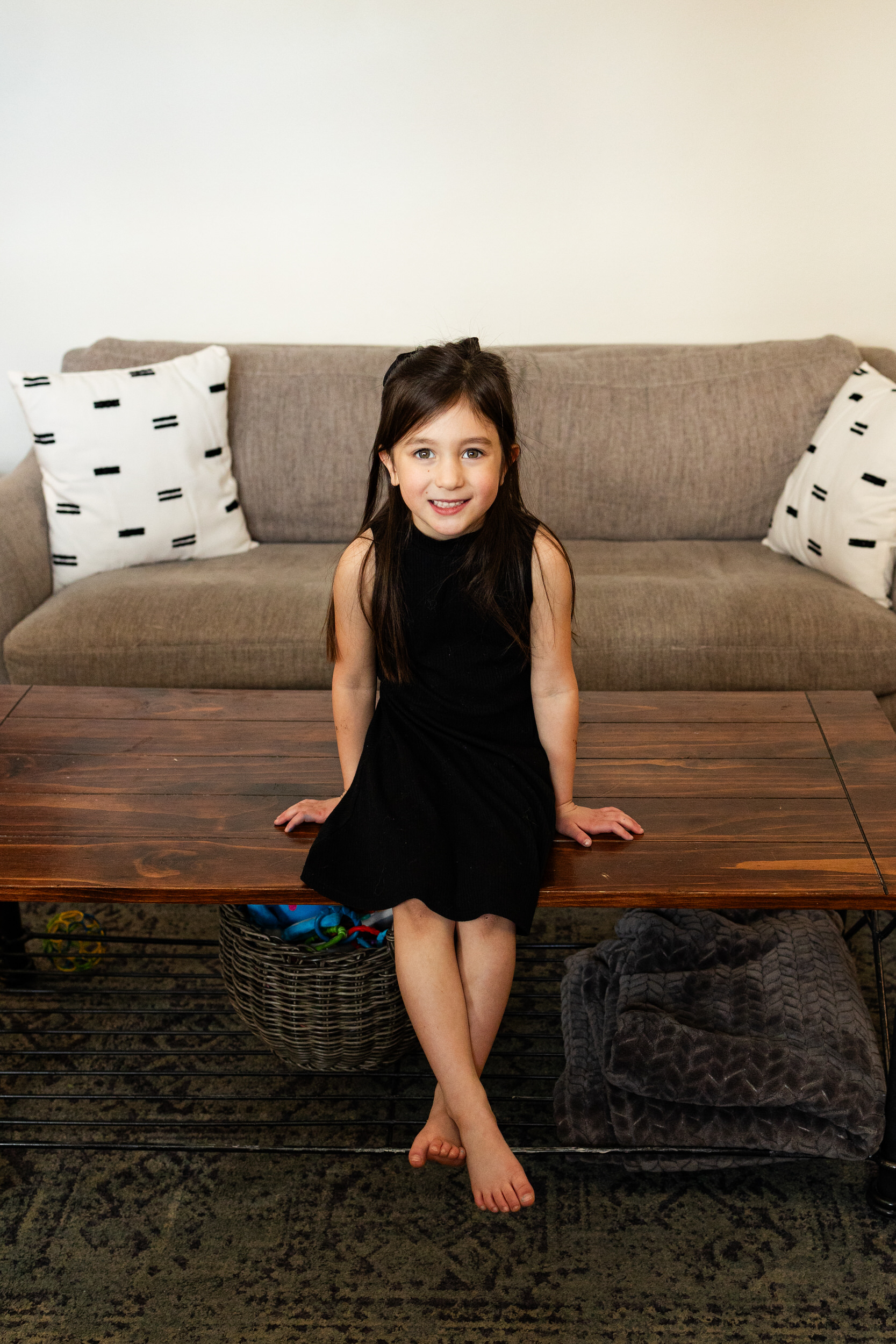 A young girl sits on a coffee table in her living room and smiles at the camera.