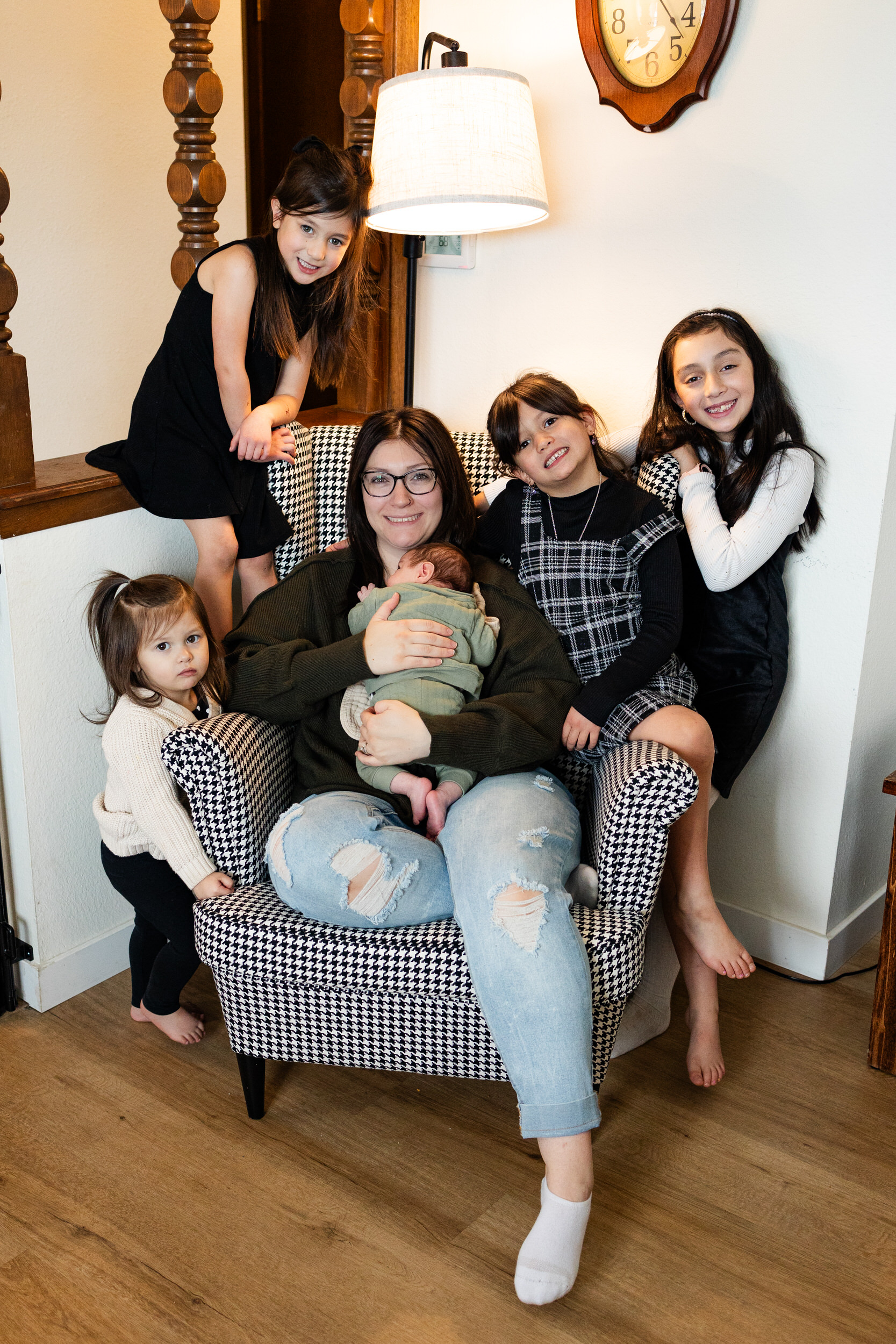 A mom is surrounded by her four young daughters as they all smile at the camera and she holds her newborn son.