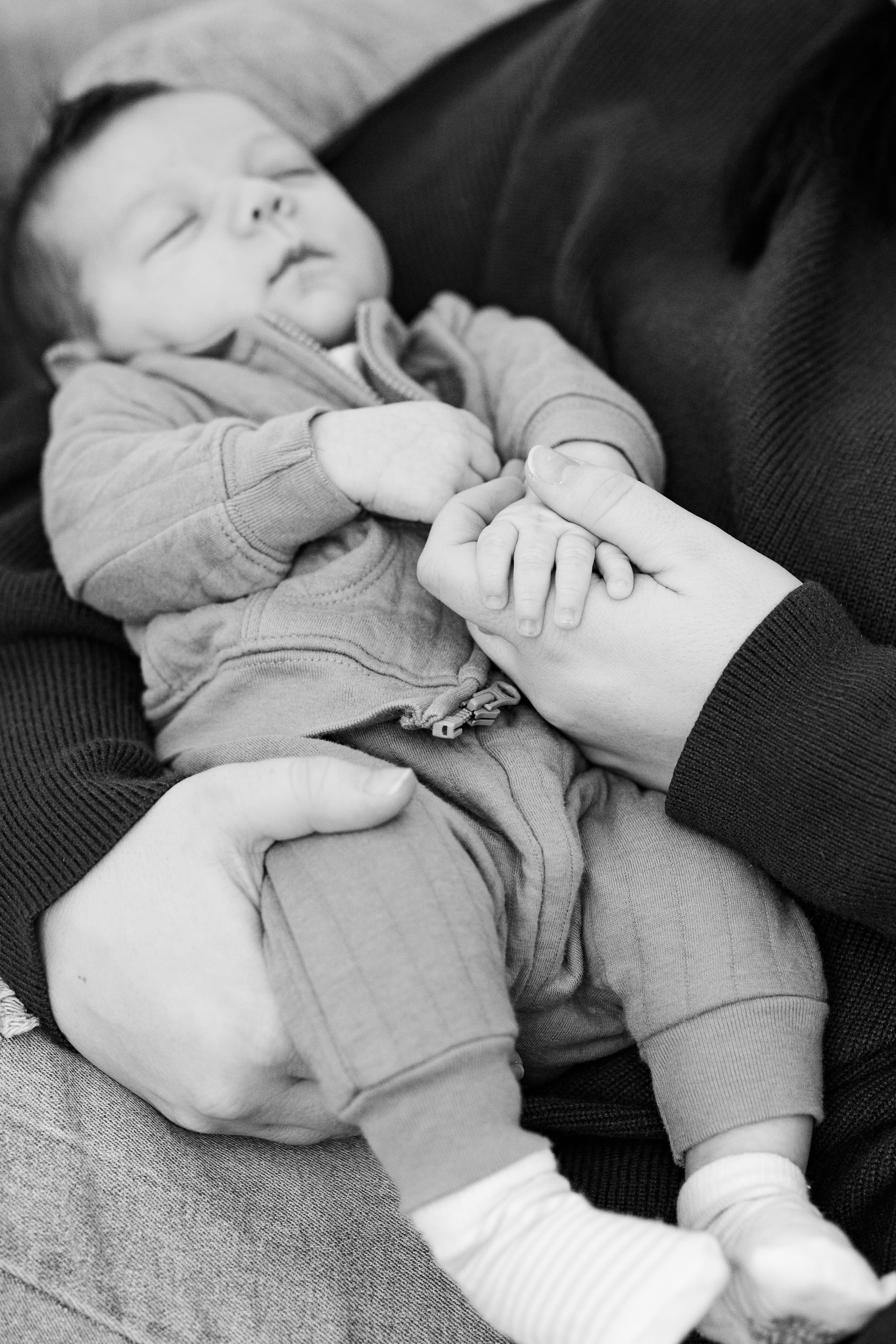 Newborn baby holds his mom's hand while he sleeps in her arms.