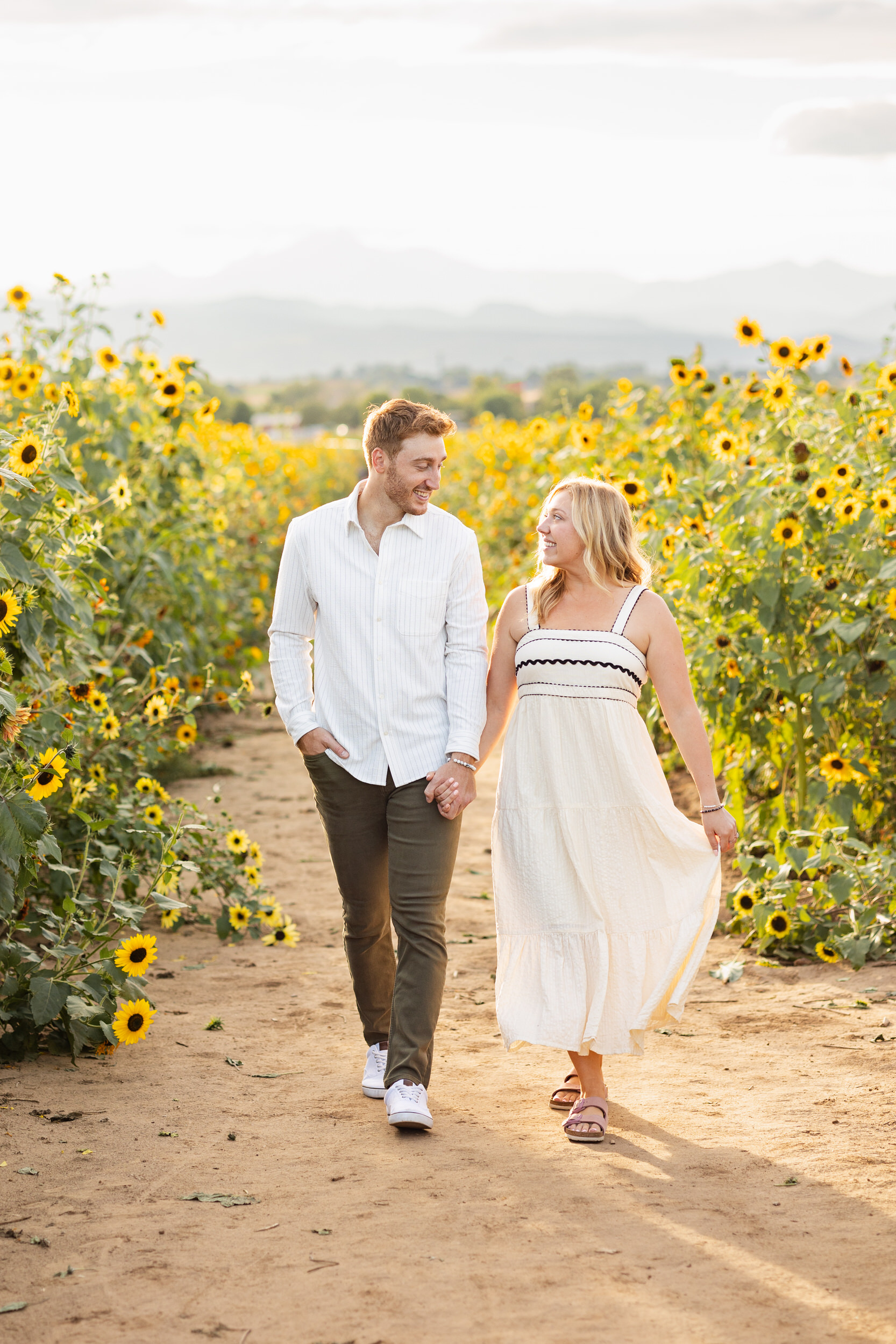 An engaged couple holds hands and smiles at each other while walking through a sunflower field.