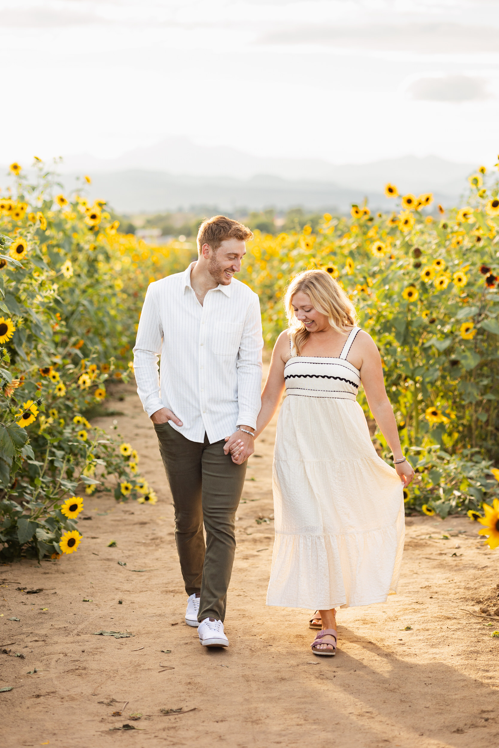 An engaged couple walks hand in hand through a sunflower field while smiling.