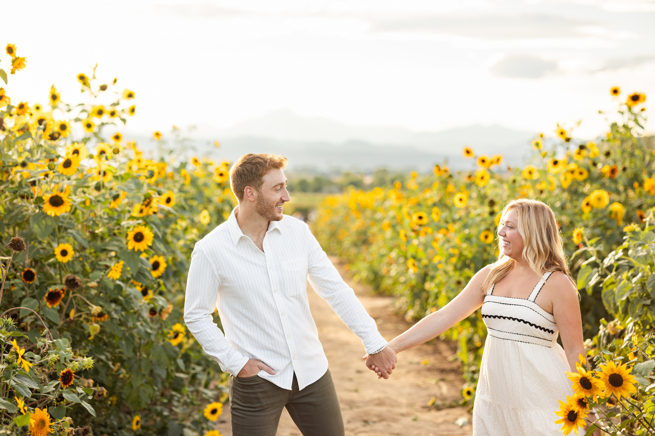 An engaged couple walks hand in hand through a sunflower field smiling at each other.