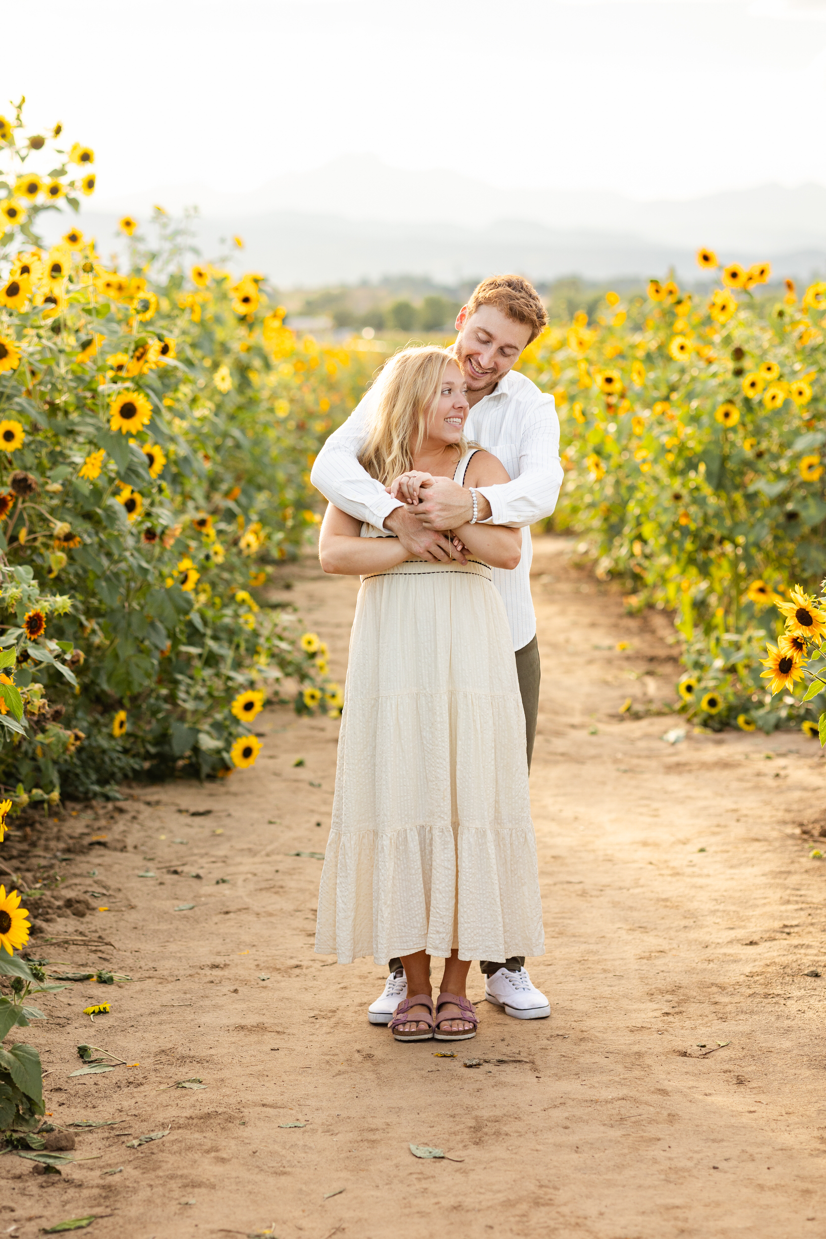 A man hugs his fiancée around her shoulders and smiles at her in a sunflower field.