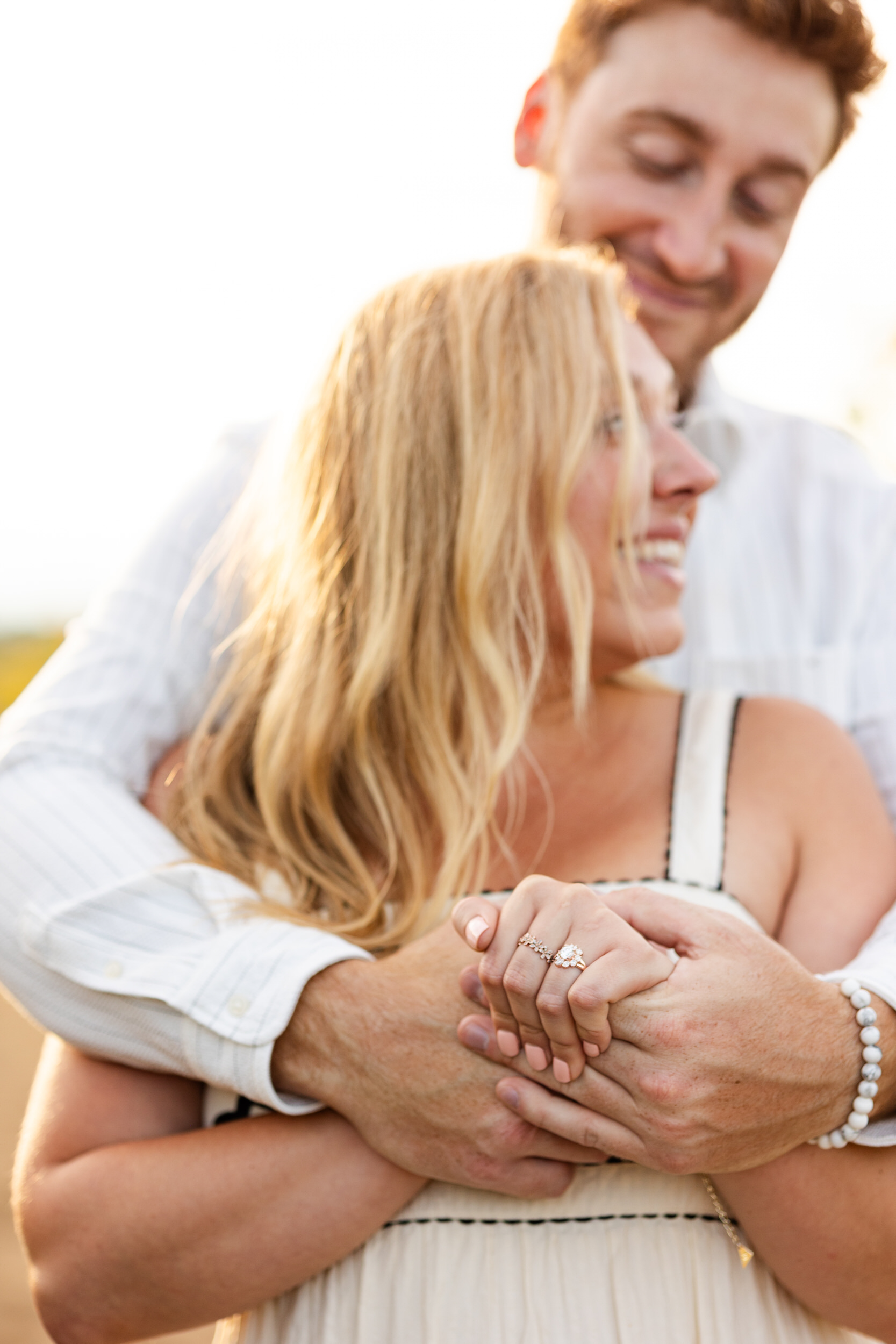 Close up of a woman's engagement ring as her fiancé hugs her around her shoulders.