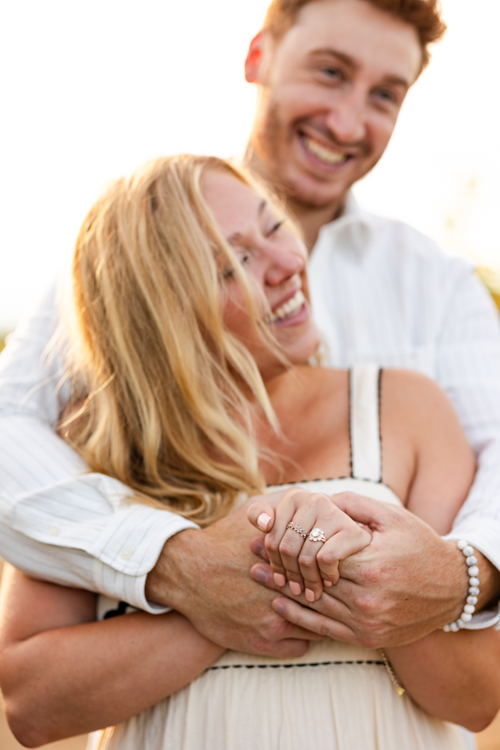 Close up of a woman's engagement ring as her fiancé hugs her around her shoulders.